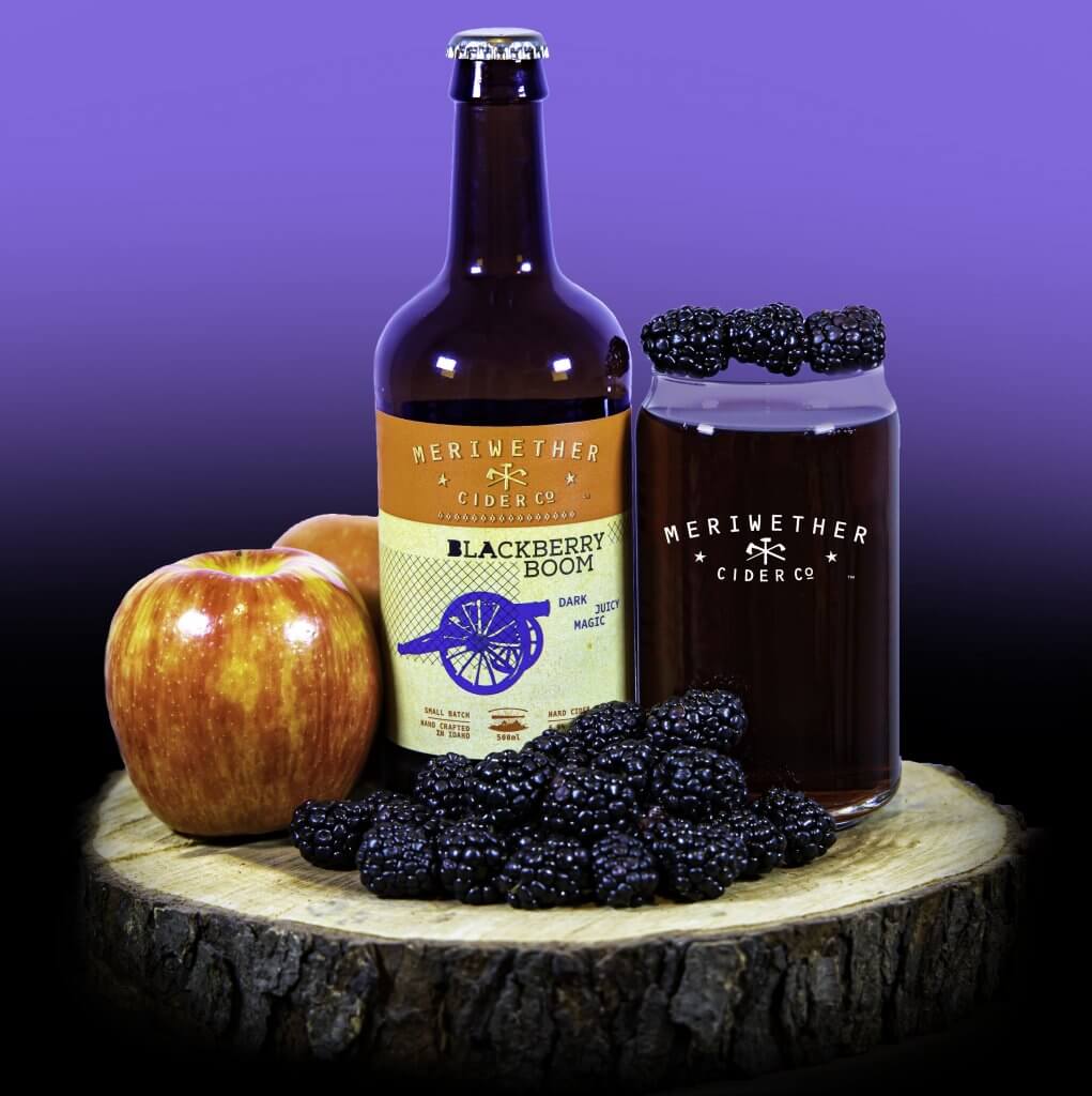 A bottle of Meriwether Cider Company's Blackberry Boom sits on a tree log surrounded by blackberries and apples.