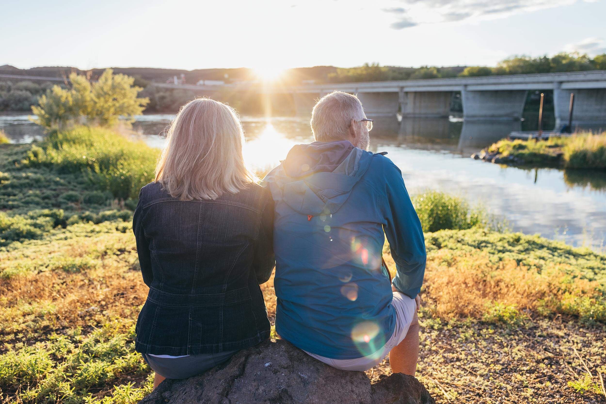 a man and woman sitting on a rock beside a river with a bridge across it