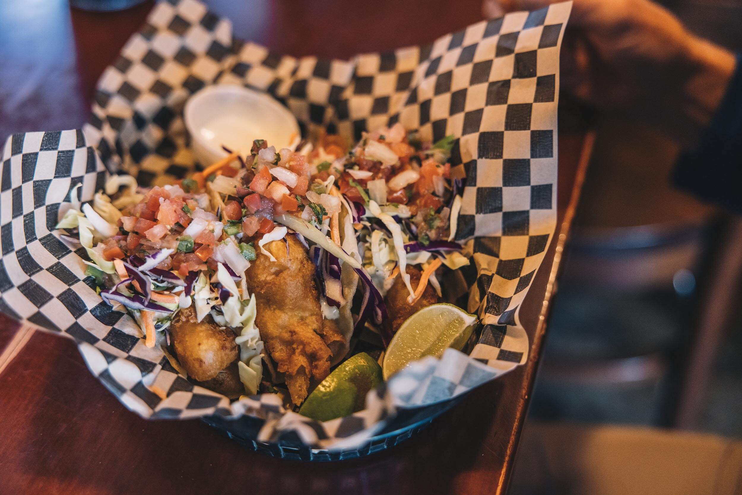 closeup of three fish tacos, two lime slices and a cup of sauce on a table