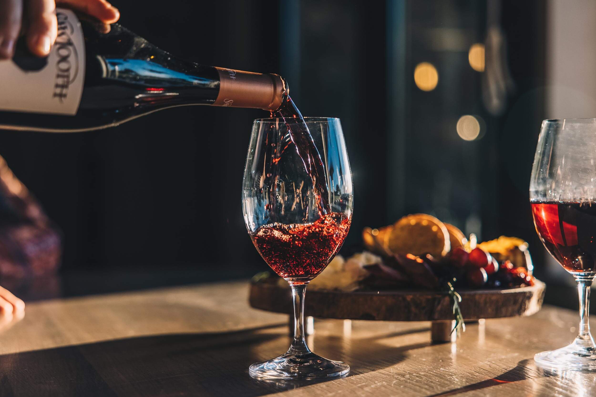 a person pouring a glass of wine with a platter of food in the background.