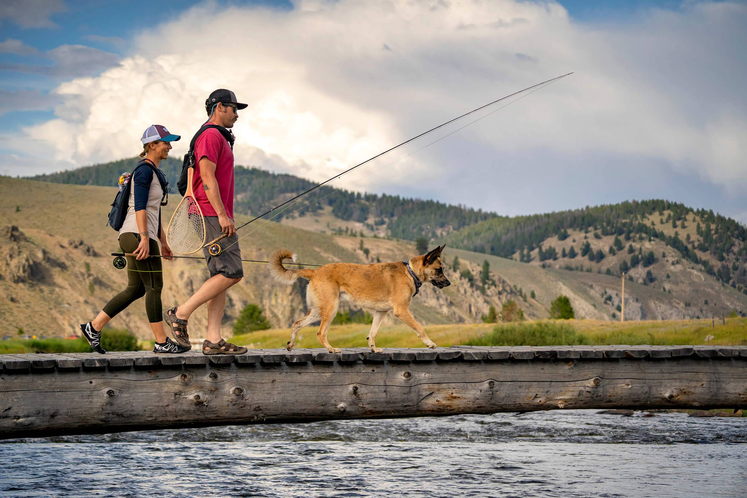 a man and a woman carrying fishing rods and walking their dog along the water with mountains in the background