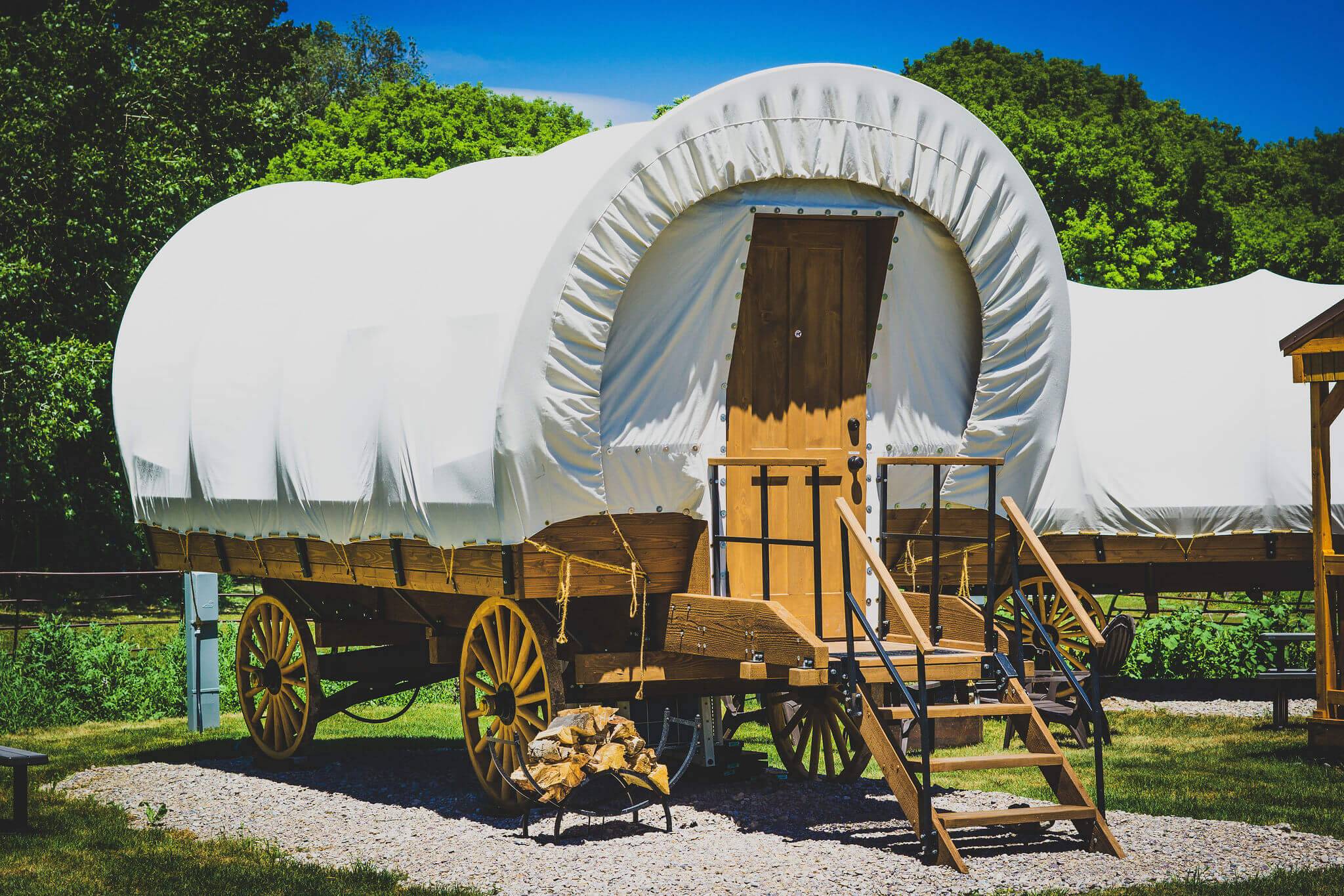 A view view of the exterior of a Conestoga Wagon, featuring wooden wheels and wooden steps that lead to the entrance outside.