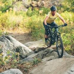 mountain biker, April Zastrow, wears a yellow shirt and rides over some rocks on her mountain bike