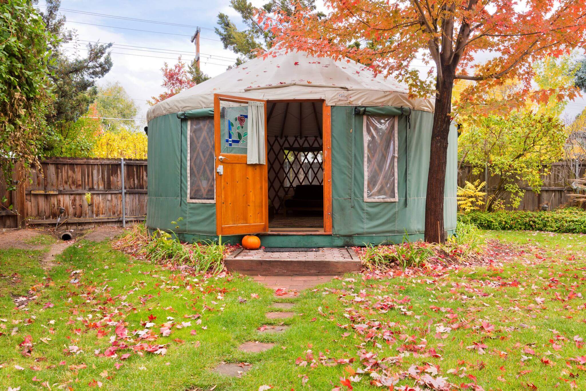 A view of the exterior of the 36th Street Urban Yurt during fall, where an orange door opens to the green building and autumn leaves line both sides of the grass around the steps that lead to the Airbnb.