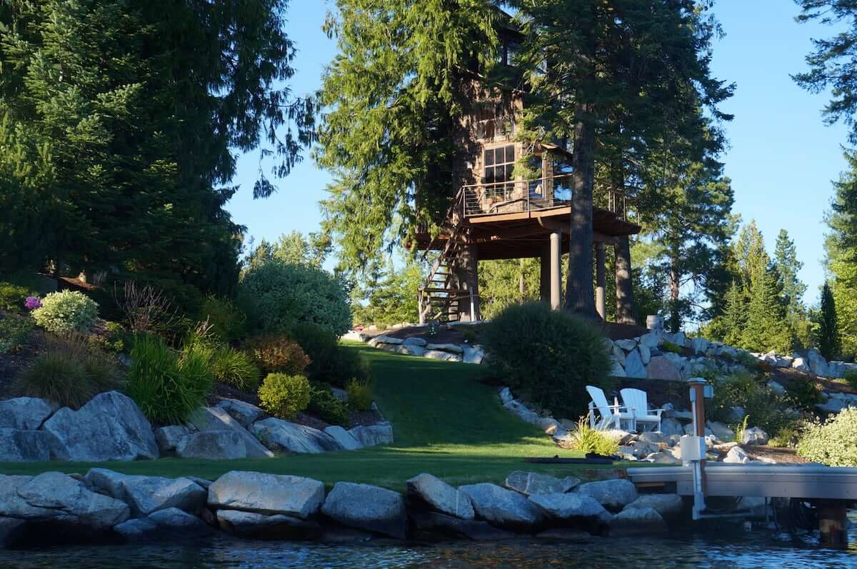 Surrounded by green grass and tall trees, this three-story treehouse sits elevated on the shore of Lake Pend Oreille in Sandpoint.