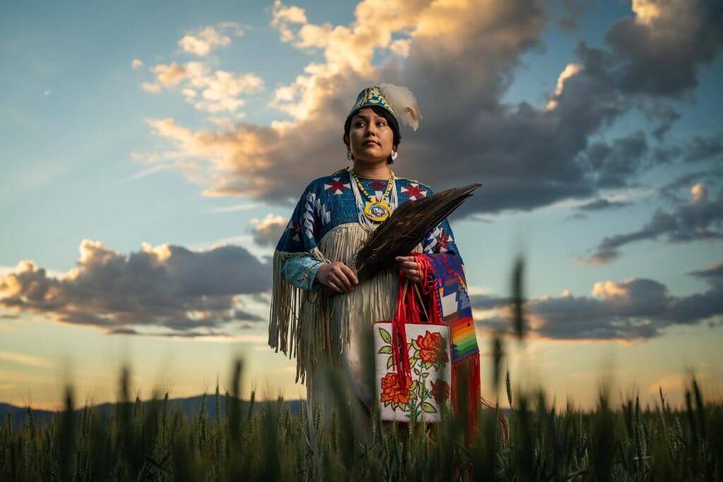 A person standing in a field holding an embroidered bag and feather fans.