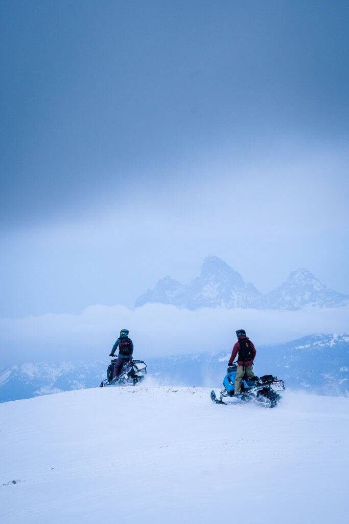 two snowmobilers look at the Grand Tetons mountains in the distance