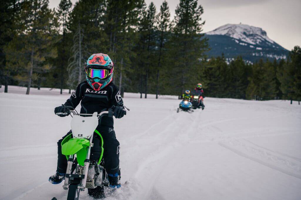 child on snobike with parent on snowmobile in background in island park