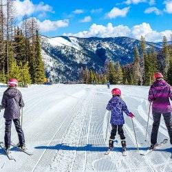 Two kids and an adult in snow gear ski a run at Lookout pass with a mountain in the distance.