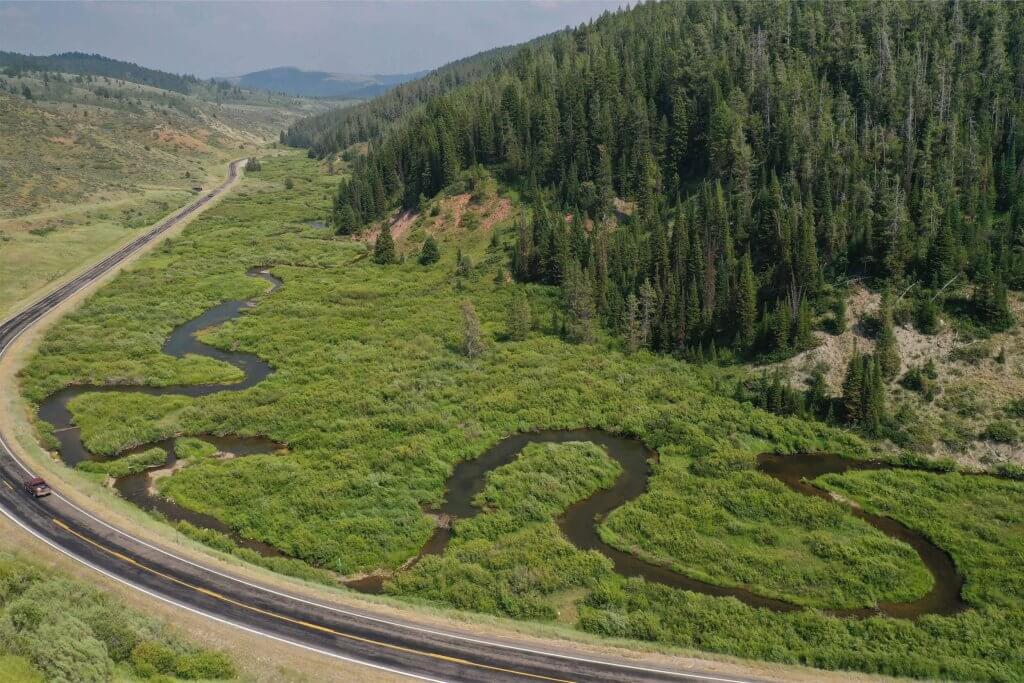 a car driving past a grassy area with a creek winding through it, beside a mountain covered with trees