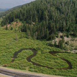 a car driving past a grassy area with a creek winding through it, beside a mountain covered with trees