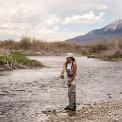 woman fly fishing in river with mountains in the background