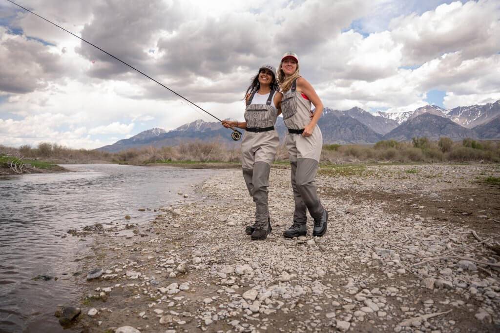 two women standing alongside river with fly fishing rods with mountains in background