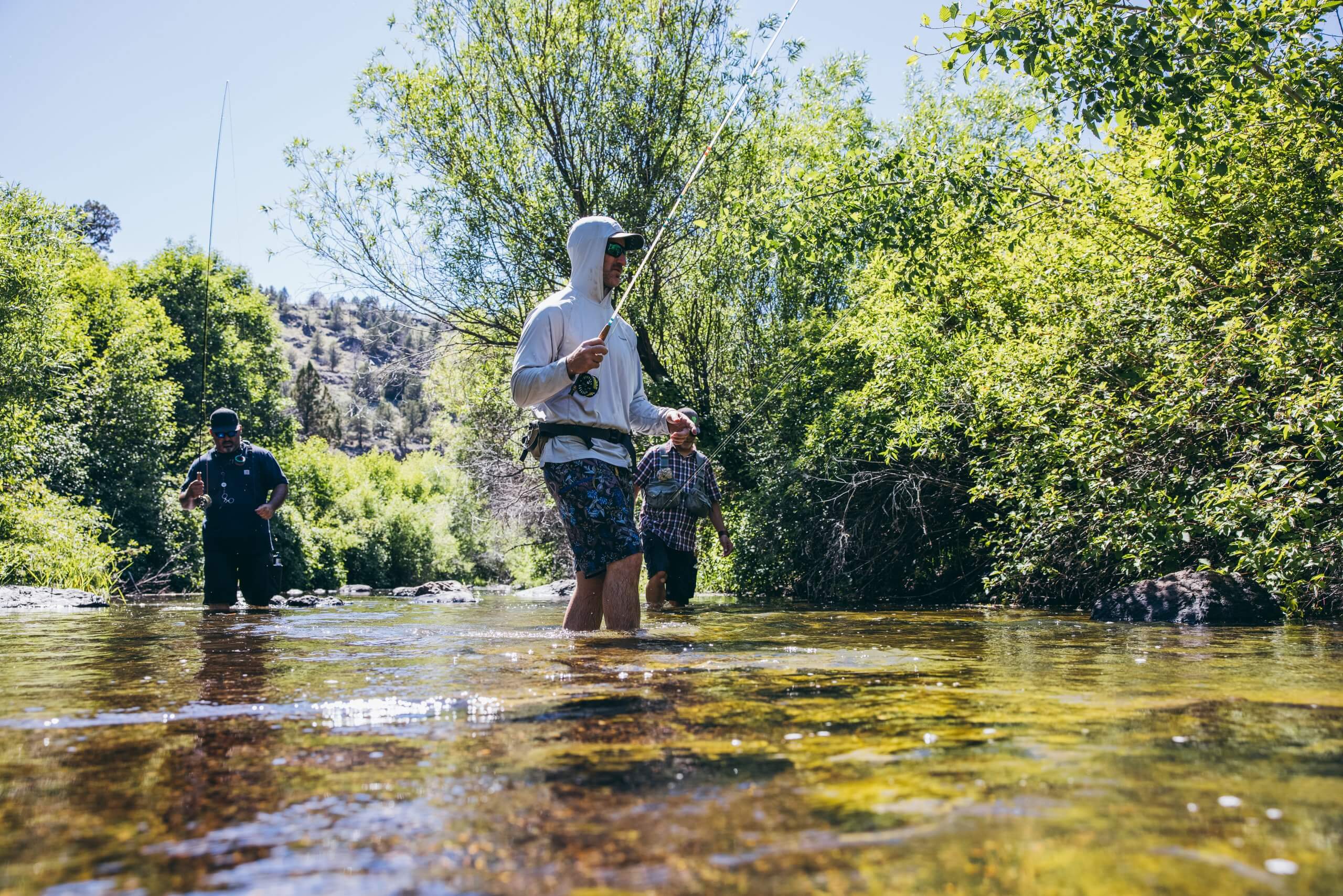 three people standing in a tree-lined creek, fishing