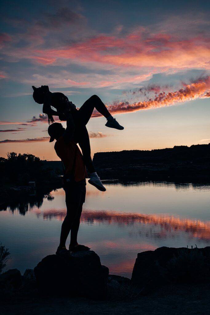 man lifting woman over his head with sunset reflecting in water in background