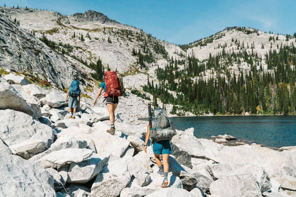 three hikers with backpackers climbing up large boulders near edge of Harrison Lake