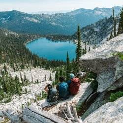 three people with backpacking gear on sitting on a rock looking at Harrison Lake far off in the distance