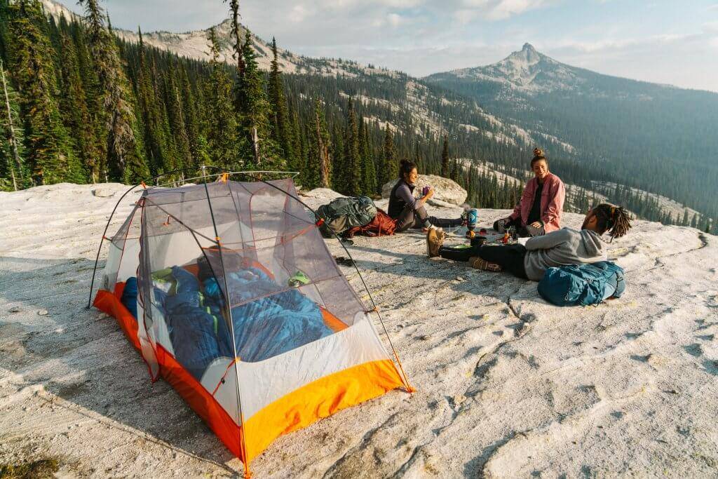 three backpackers laying on large rock slab next to a tent with mountains and trees in the distance