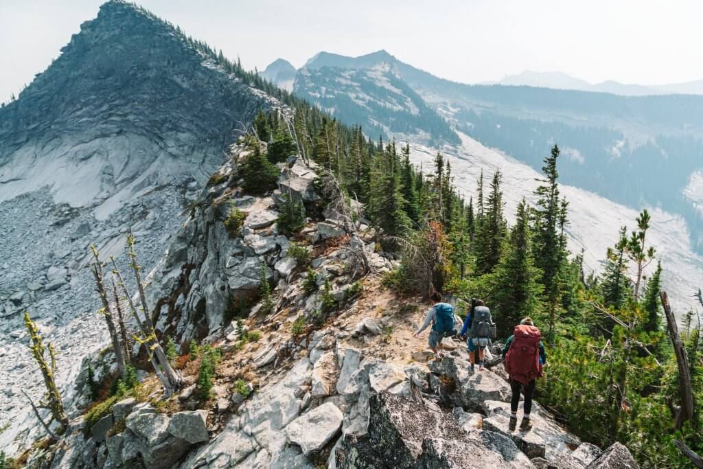 three backpackers on rocky mountain ridgeline with some trees on each side and more mountains in the distance