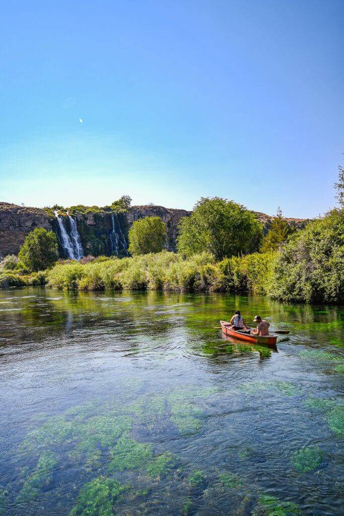 two people in a canoe on clear blue water looking at a waterfall in the distance