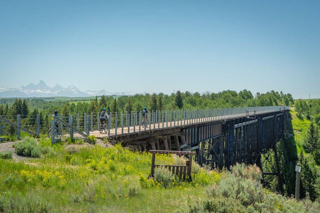 three bike riders beginning to cross train trestle style bridge with Teton Mountain Range in background