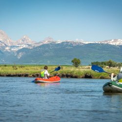 Two people in inflatable kayaks paddling the Teton River with Teton Mountain Range in the background.