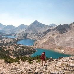 female backpacker looking over three blue alpine lakes while standing on boulder field.