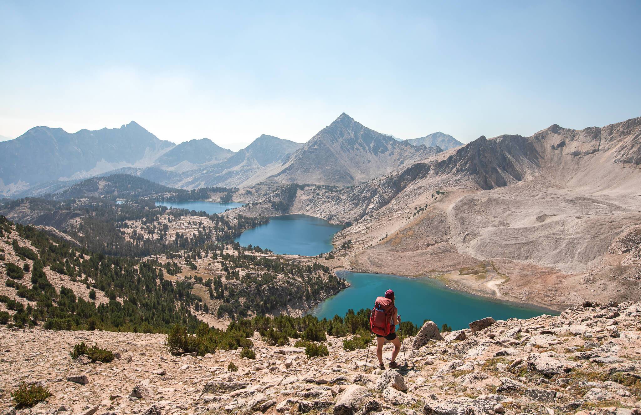 female backpacker looking over three blue alpine lakes while standing on boulder field.