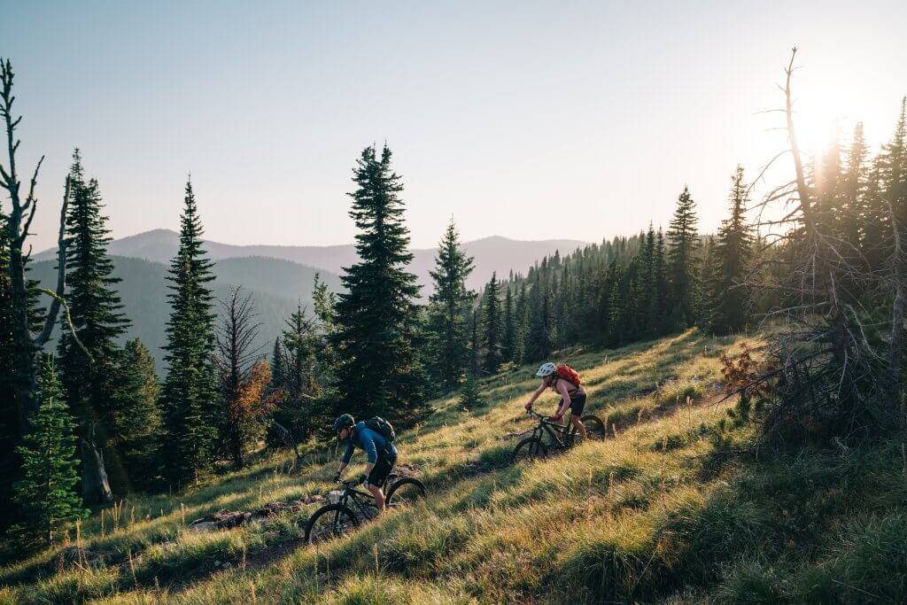 man and woman wearing helmets mountain biking on trail with tall grasses and dense forest in background