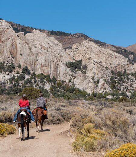 two horseback riders on dirt trail riding away with rugged rock formations in the distance at Castle Rocks State Park.