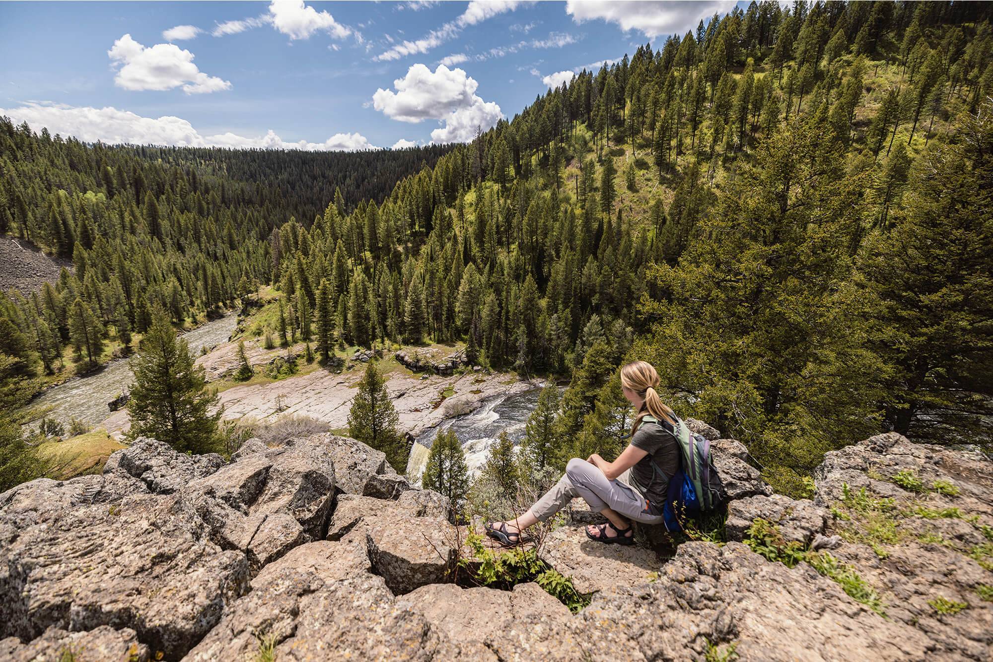 A woman sits on the edge of a cliff, overlooking a river.