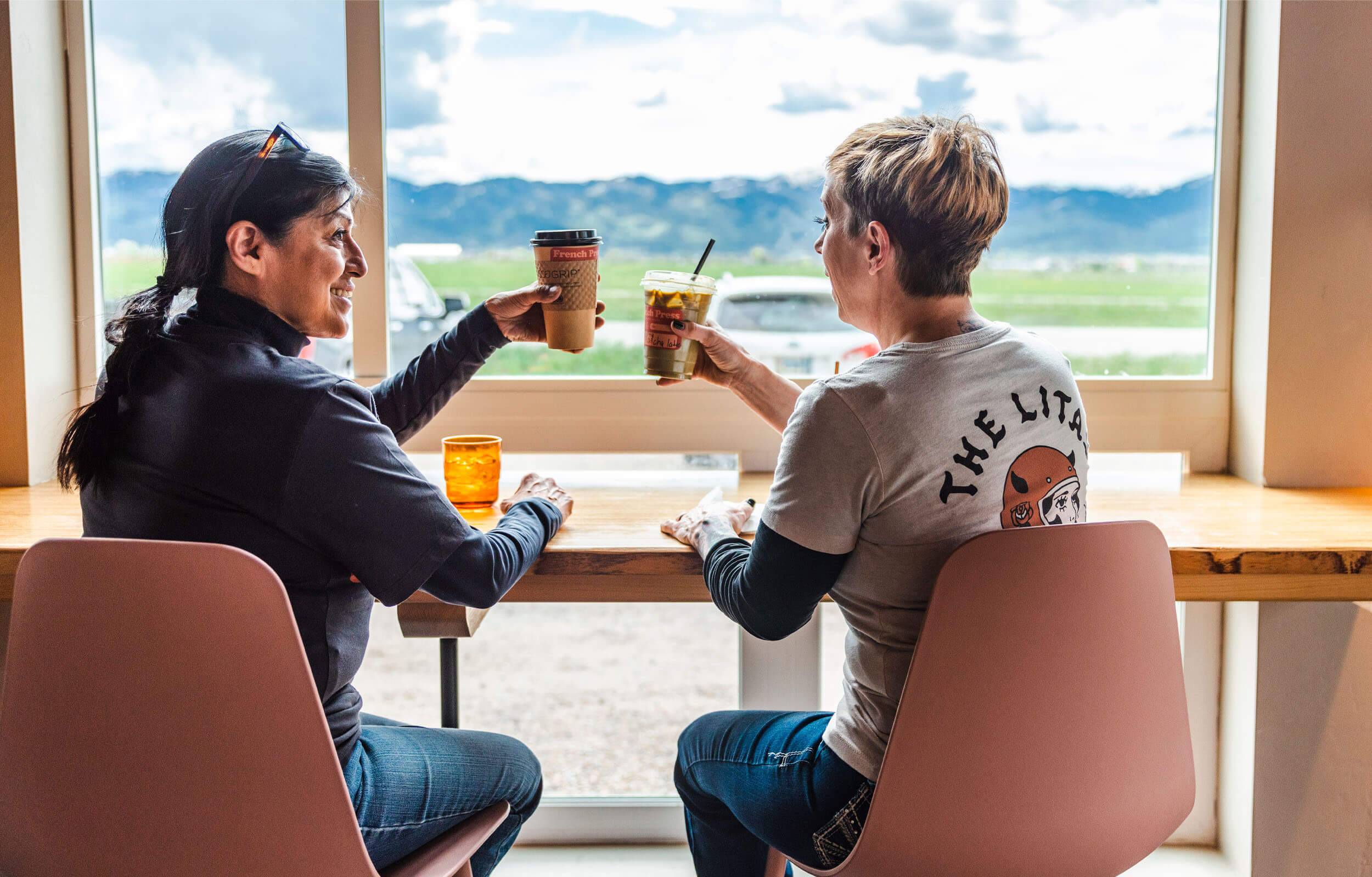 Two women cheers their coffees while sitting next to a window.