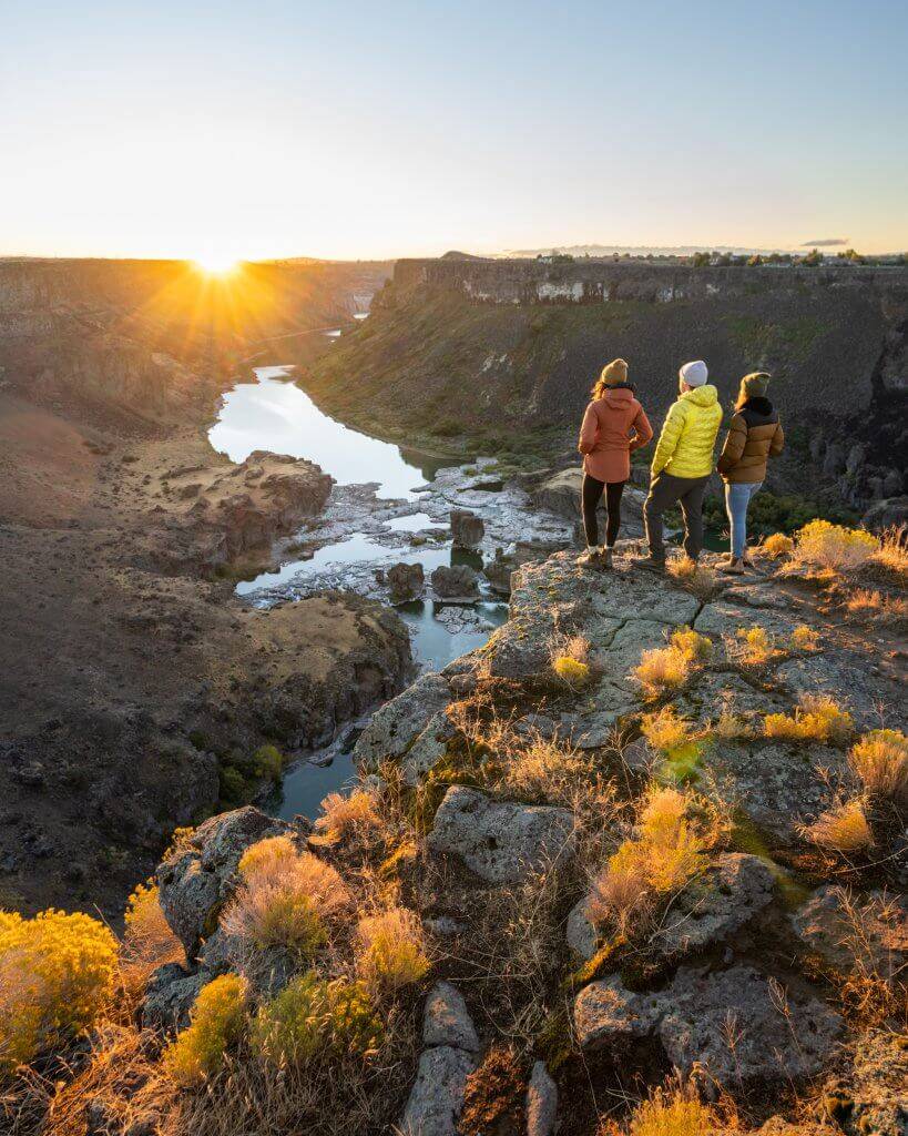 three people is mid-weight jackets standing on rocky canyon edge looking at water in the Snake River and Pillar Falls below