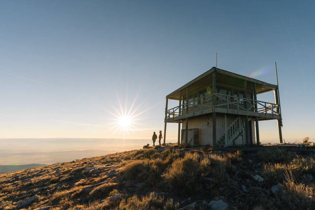 two people and a dog and a fire lookout are silhouetted by the sun