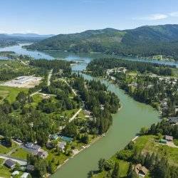 Aerial view of a river running through the town of Priest River, with tree covered mountains in the distance.