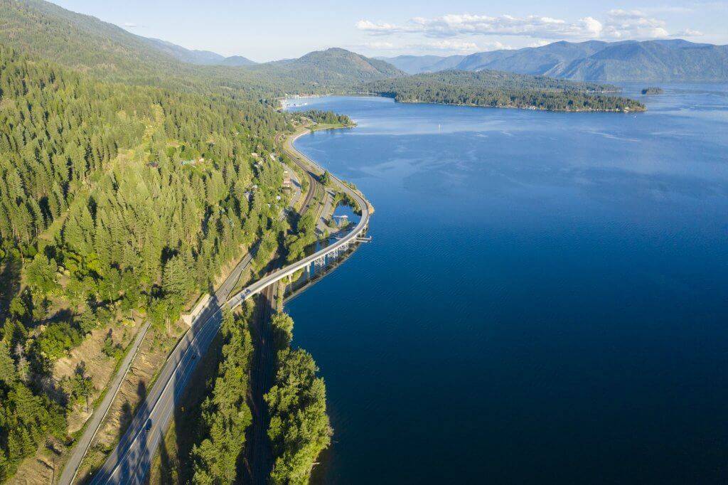 An aerial view of the Pend Oreille Scenic Byway winding between a forest and Lake Pend Oreille.