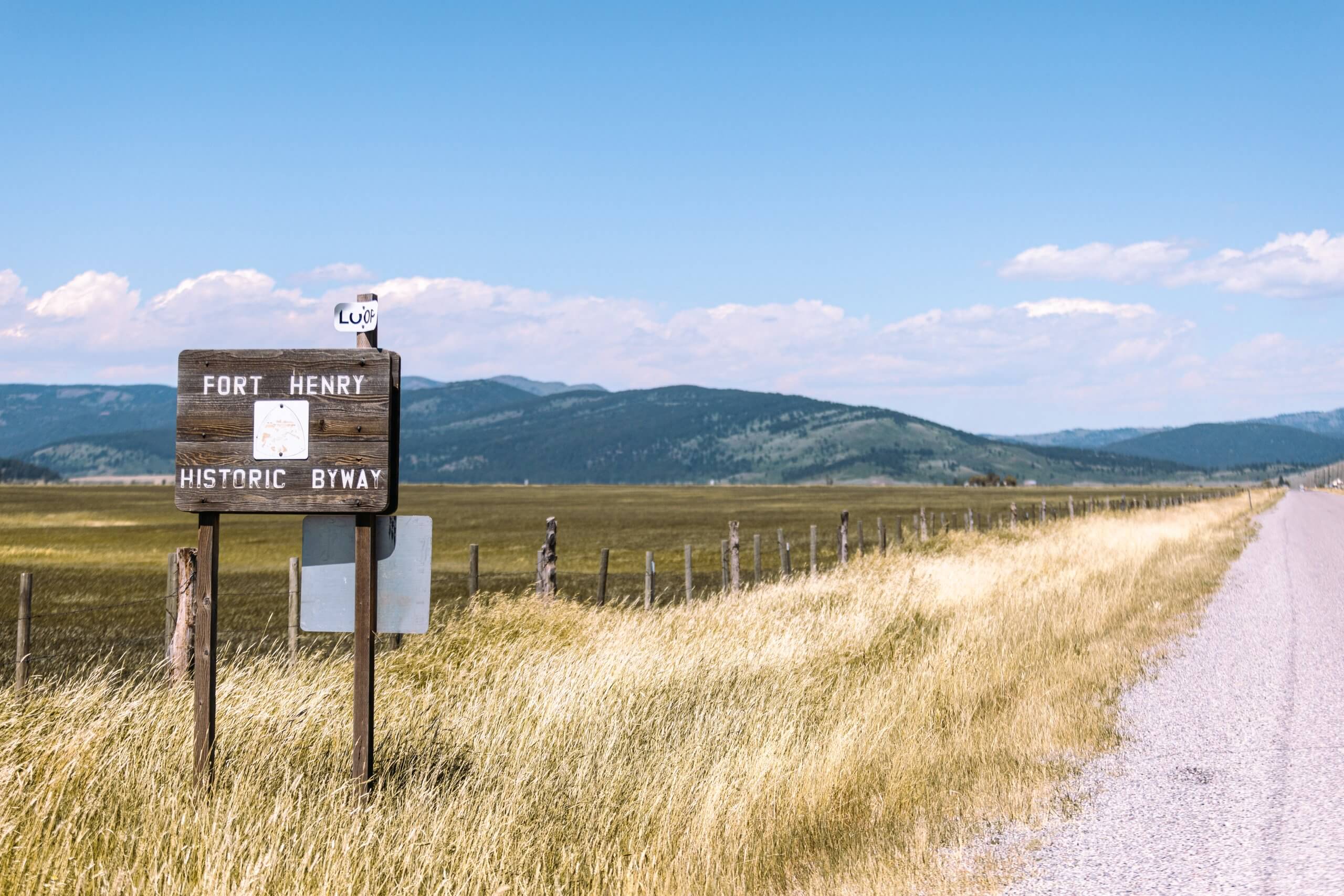 a wooden road sign that reads "fort henry historic byway", beside a sprawling field along a gravel road