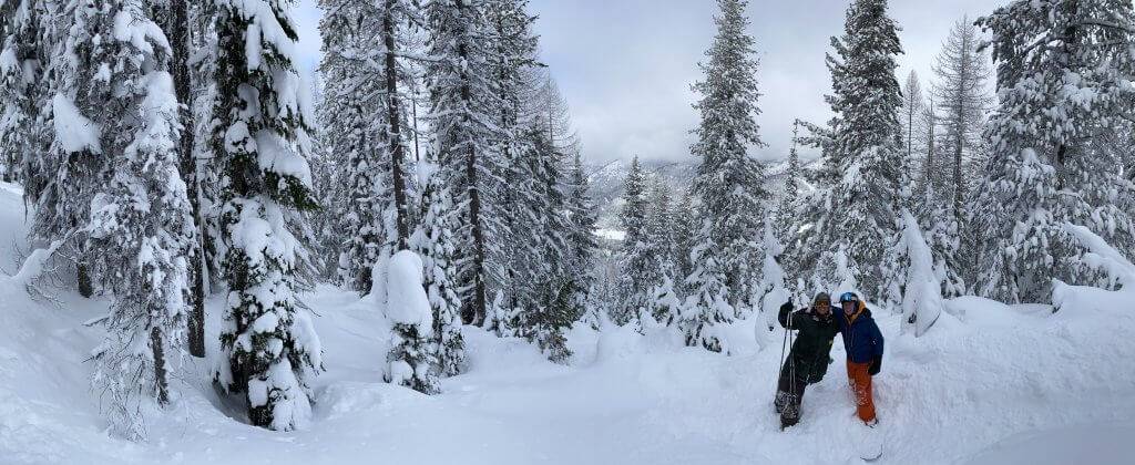 two skiers standing next to each other surrounding by snow with snow covered trees in the background