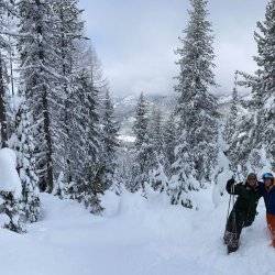 Two skiers in snow gear standing next to each other outside surrounded by deep drifts and snow-covered trees.