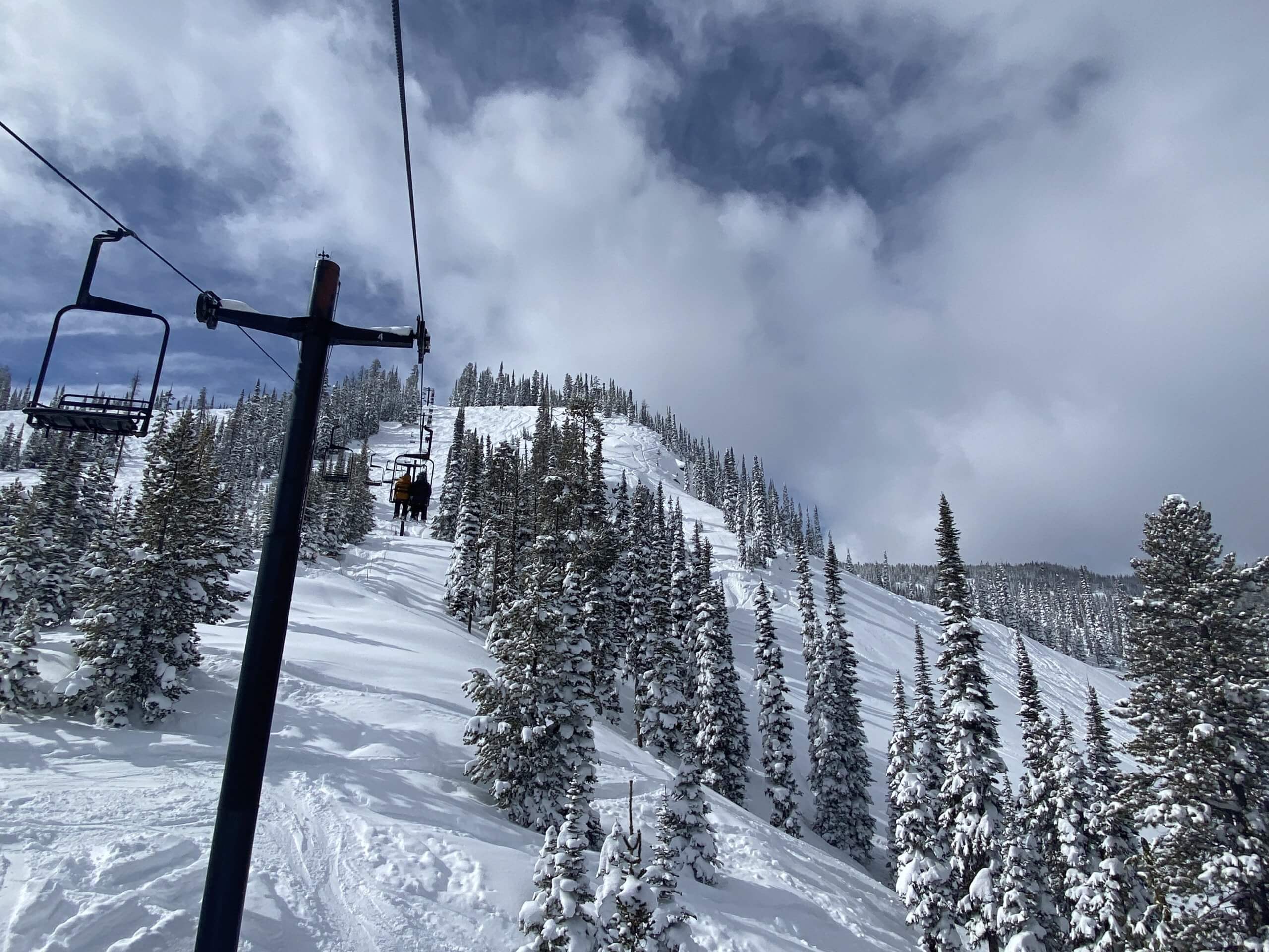 looking up a snowy mountain from the seat of t chairlift with snow on ground and skiers on the lift seat in front