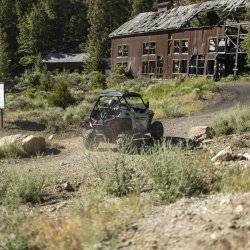 A person drives a UTV down a dirt road toward a historic building on the Mackay Mine Hill Tour.