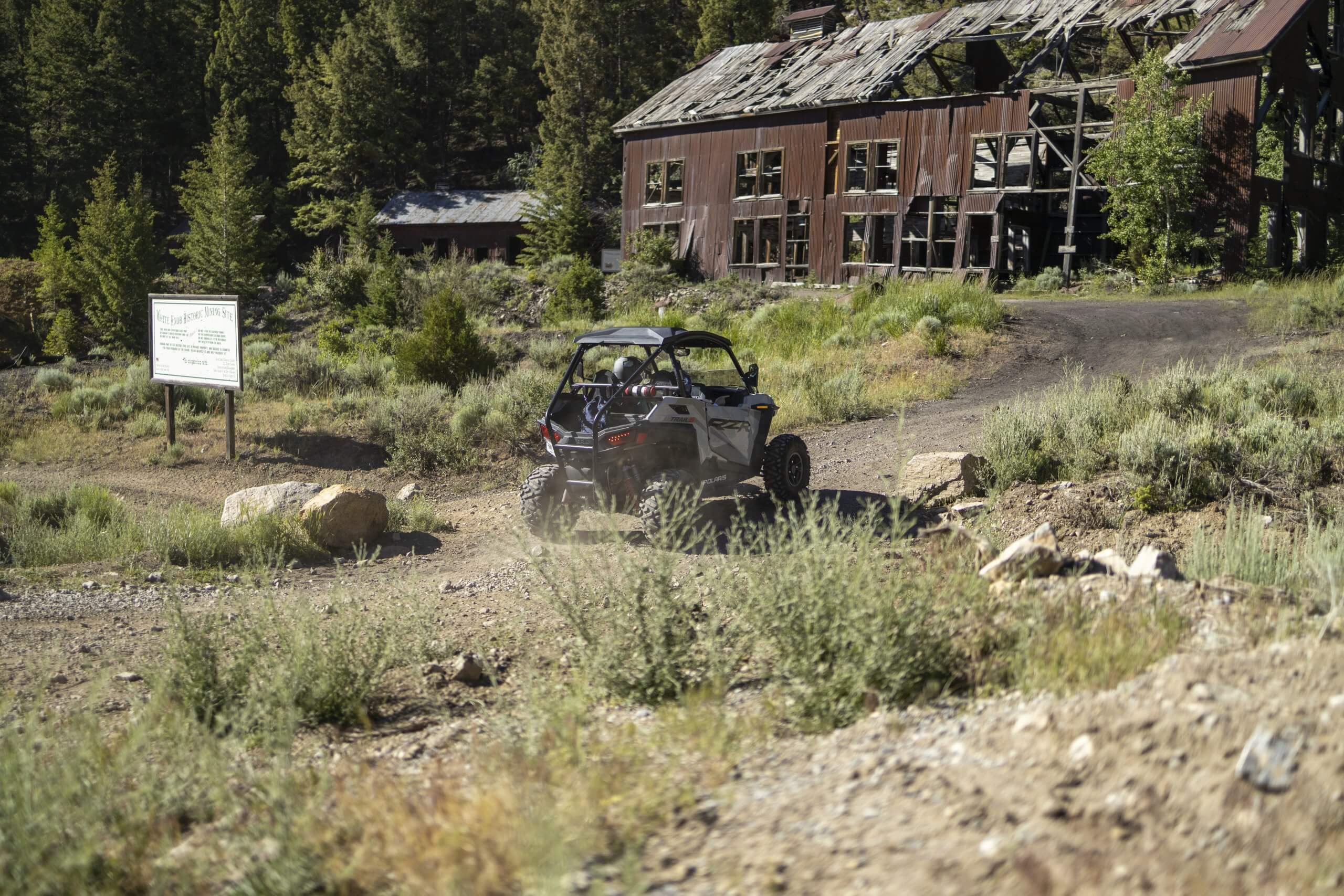 A person drives a UTV down a dirt road toward a historic building on the Mackay Mine Hill Tour.