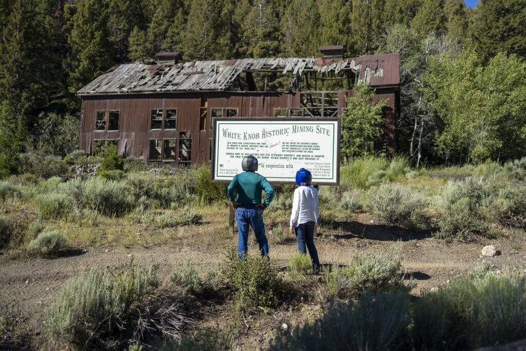 Two people in helmets stand in front of the White Knob Historic Mining site and sign. 