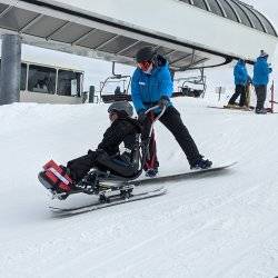 A ski instructor on a snowboard behind a skier in a sit ski helping them down the slope near the lift.