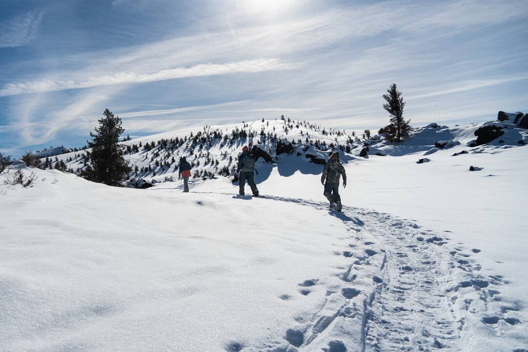 thee people in heavy winter coats are snowshoeing in a line on top of a snowy path at craters of the moon