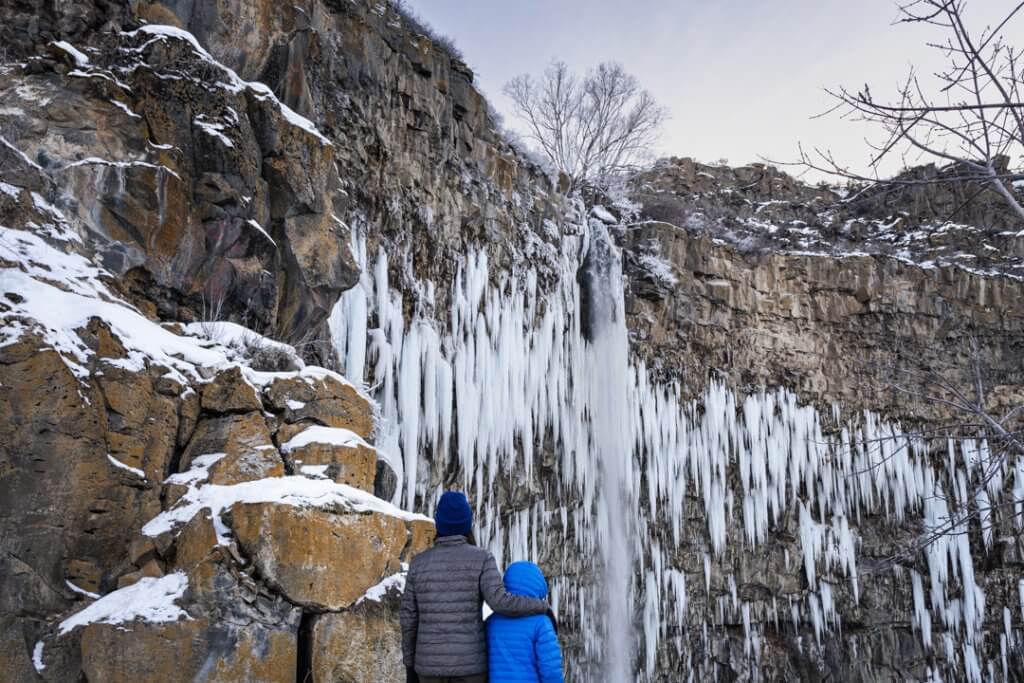 two people in winter coats standing near the base of a frozen waterfall looking up