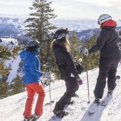 three skiers stand next to each other on snowy mountainside looking at snowy mountains in the distance