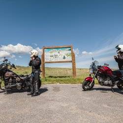 two motorcycle riders in helmets standing next to Teton Scenic Byway signs with blue skies in the background