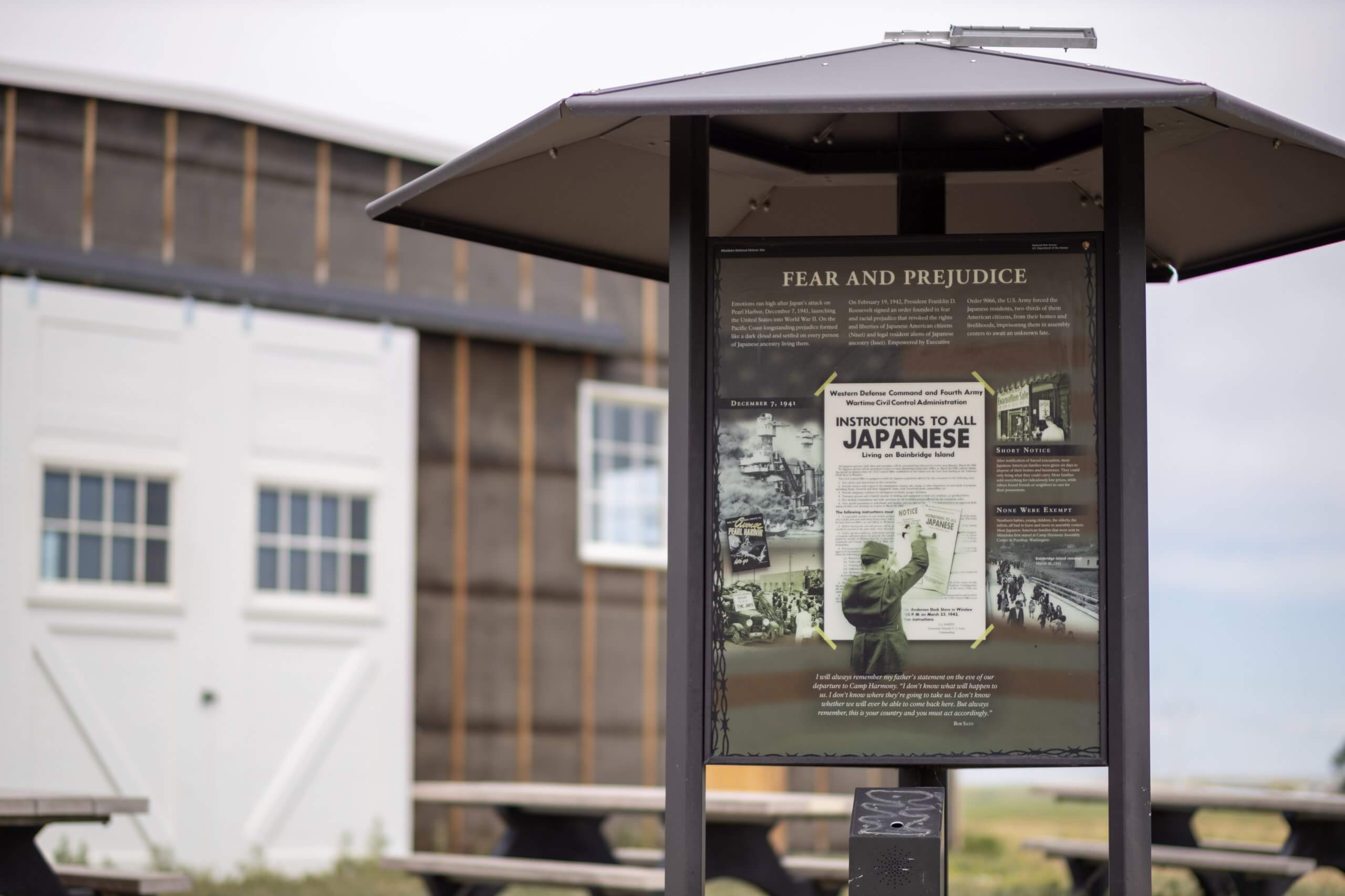 Close-up of a sign containing historical information in front of a building and picnic tables at Minidoka National Historic Site.