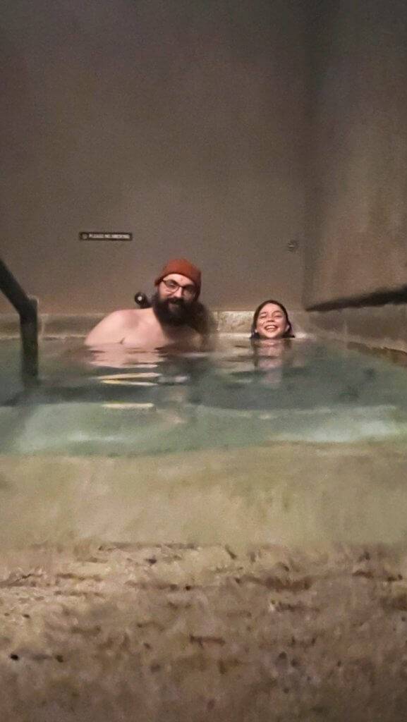 a father and daughter sitting in a private hot pool with water up to their shoulders, smiling